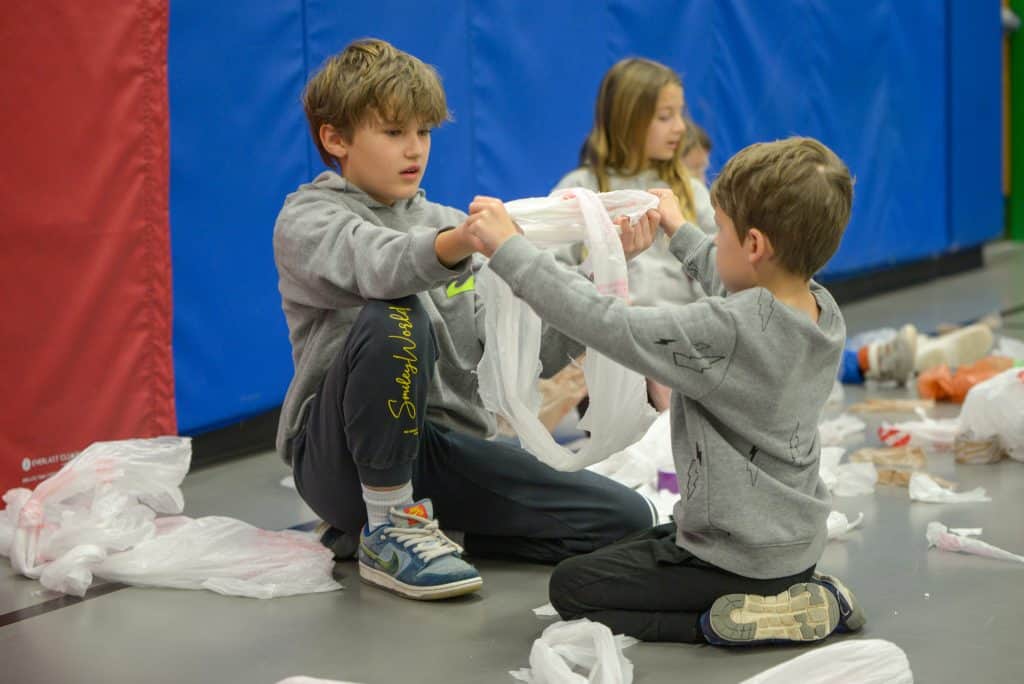  A 5th grader and 1st grader working on a craft project making jump ropes out of plastic bags. They are seated on the floor facing each other and their arms are extended as they hold onto the bags between them. 