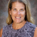 A woman with long, wavy hair smiles at the camera, wearing a sleeveless, patterned top against a textured background.