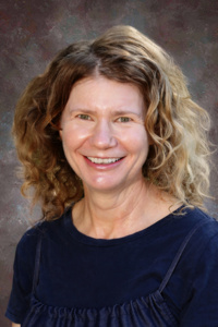 Smiling woman with curly hair, wearing a dark blue top, against a textured gray background.