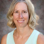 A smiling woman with shoulder-length blonde hair wearing a light blue sleeveless top, set against a textured background.