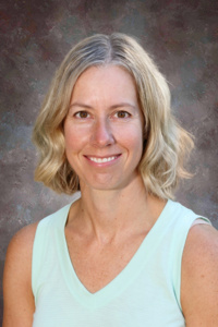 A smiling woman with shoulder-length blonde hair wearing a light blue sleeveless top, set against a textured background.