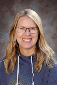 Smiling woman with long blonde hair, wearing glasses and a blue hoodie, set against a blurred background.