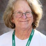 Smiling woman with glasses and curly hair, wearing a white shirt and a green lanyard from a school.