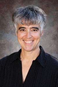 Smiling person with short, wavy gray hair wearing a black shirt, against a soft, muted background.