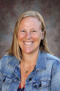 Smiling woman with shoulder-length blonde hair wearing a denim jacket, set against a neutral background.