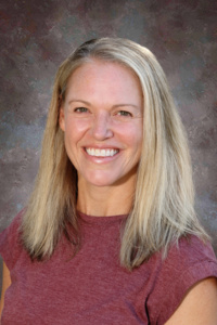 Smiling woman with long, straight blonde hair wearing a maroon shirt against a textured gray background.