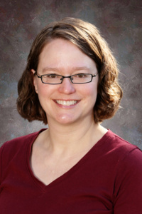 Smiling woman with shoulder-length brown hair, wearing glasses and a maroon shirt, against a neutral background.