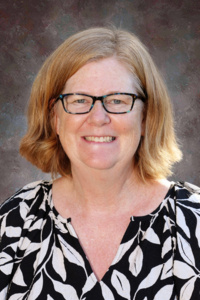 A smiling woman with shoulder-length hair, wearing glasses and a black-and-white patterned blouse, poses against a neutral background.