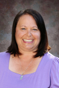 Smiling woman with shoulder-length dark hair, wearing a purple top, against a textured gray background.