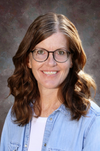 A woman with wavy brown hair and glasses smiles, wearing a light blue shirt against a textured gray background.