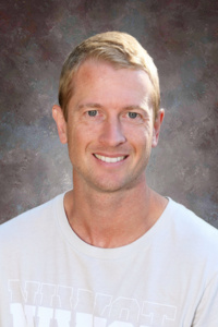 Smiling man with short blonde hair, wearing a light-colored t-shirt, set against a neutral background.