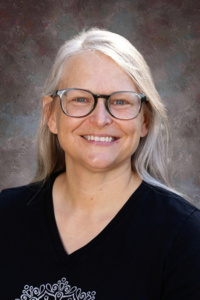 Smiling woman with long, light gray hair and glasses, wearing a black shirt with a decorative design.