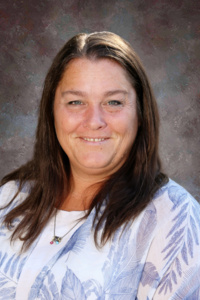 A smiling woman with long brown hair, wearing a white shirt with blue leaf patterns, against a textured background.
