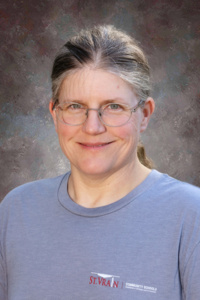 A smiling middle-aged woman with glasses and long hair, wearing a gray t-shirt, against a textured background.
