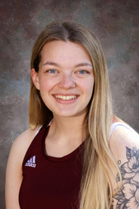 Young woman with long hair and a tattoo, wearing a maroon athletic top, smiling against a textured background.