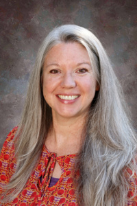 Smiling woman with long gray hair, wearing a colorful patterned blouse, against a softly blurred background.