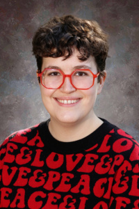 A smiling person with short curly hair and red glasses wears a black sweater with "LOVE" and "PEACE" printed on it.