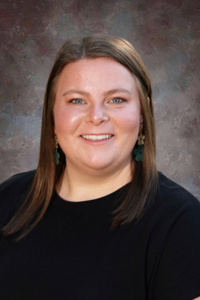 A smiling woman with long brown hair, wearing a black shirt and green earrings, against a textured background.
