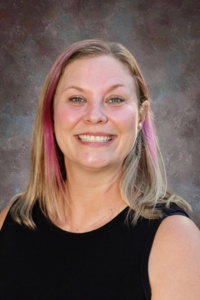 A smiling woman with shoulder-length hair, featuring pink highlights, wearing a black sleeveless top against a textured background.