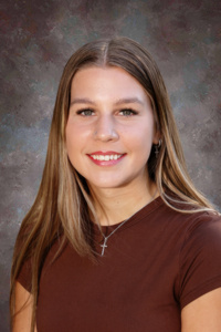A smiling young woman with long brown hair, wearing a brown shirt and a cross necklace, poses for a portrait.