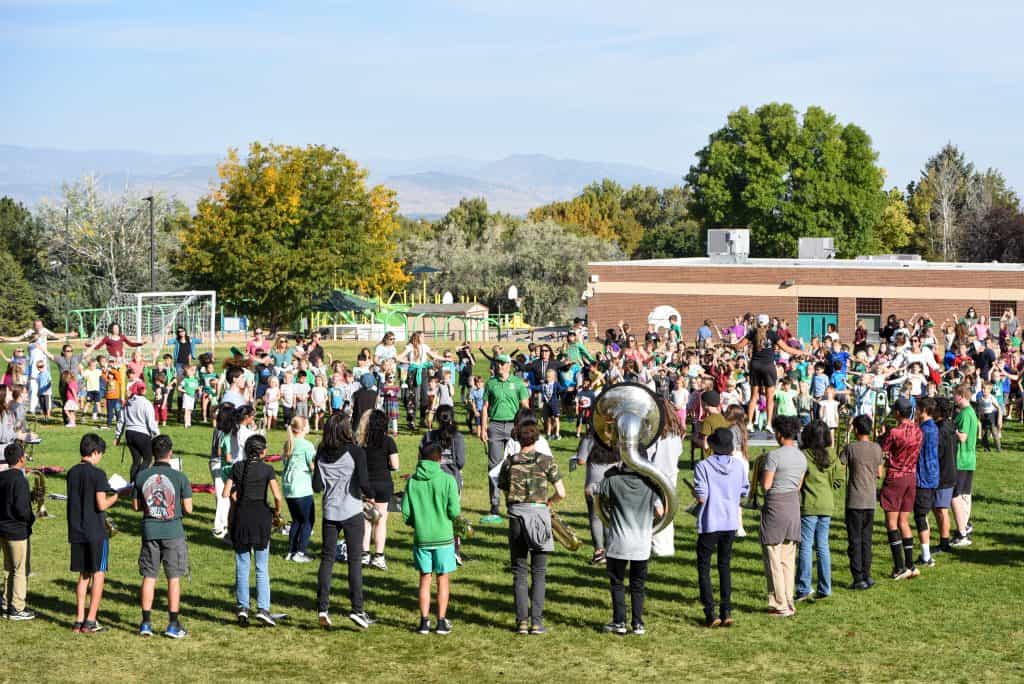 Photo from afar of the Niwot High School band standing in a large circle with their backs to the camera. They are facing the elementary students at in front of them who are dancing to their music on the grass field outside of the school. 