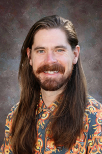 A man with long hair and a beard smiles, wearing a colorful patterned shirt against a neutral background.