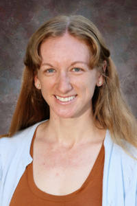 A smiling woman with long, wavy hair, wearing a light blue cardigan over a brown top, against a textured background.