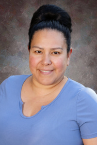 A smiling woman with dark hair styled in a bun, wearing a light blue shirt, against a textured background.