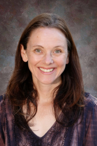 Smiling woman with long brown hair, wearing a dark purple top, against a textured gray background.