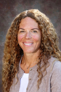Smiling woman with long, curly hair wearing a light-colored top and cardigan, set against a neutral background.