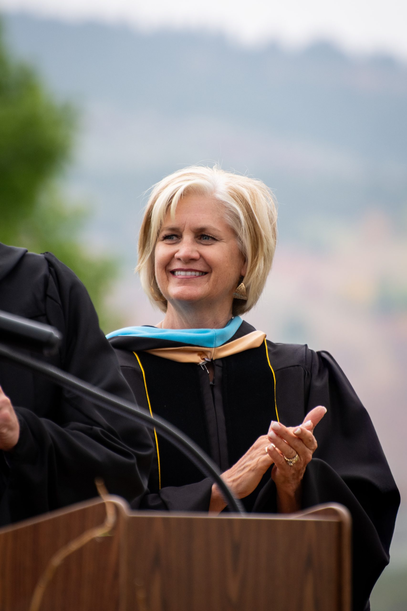 Smiling woman in graduation gown and hood claps at a podium with blurred outdoor background.