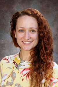 A smiling woman with long, curly reddish-brown hair, wearing a colorful floral shirt, against a neutral background.