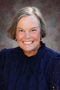 Smiling woman with short gray hair, wearing a navy blue blouse, against a textured neutral background.