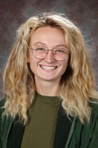 Smiling young woman with curly blonde hair, wearing glasses and a green shirt, against a textured background.
