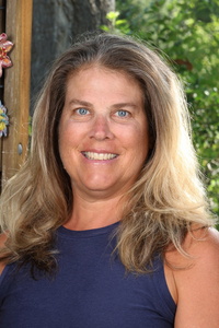 Smiling woman with long, wavy hair, wearing a sleeveless top, stands outdoors with greenery in the background.
