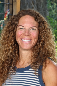 Smiling woman with curly hair wearing a striped tank top, standing outdoors with greenery in the background.