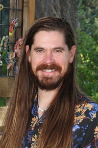 A man with long hair and a beard smiles, wearing a floral shirt, with greenery in the background.