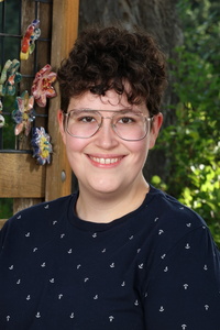 A person with curly hair and glasses smiles in front of a wooden structure adorned with colorful decorations.