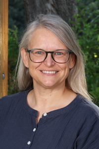 Smiling woman with long, light gray hair and glasses, wearing a dark shirt, standing outdoors with greenery in the background.