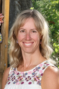 Smiling woman with long, wavy hair wearing a floral-patterned top, standing outdoors with greenery in the background.