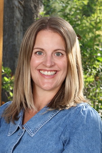Smiling woman with long, straight hair wearing a denim shirt, standing outdoors against a green background.