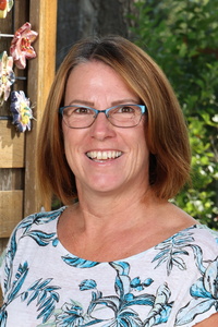 Smiling woman with shoulder-length brown hair, wearing glasses and a floral top, standing outdoors.