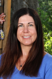 A woman with long dark hair smiles, wearing a blue shirt, with greenery and colorful decorations in the background.