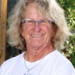 Smiling older woman with curly hair and glasses, wearing a white shirt, surrounded by greenery.