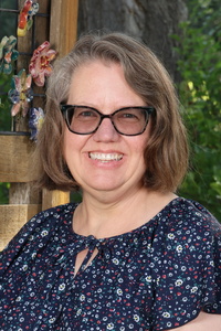 Smiling woman with shoulder-length hair, wearing glasses and a floral blouse, posed outdoors against a wooden backdrop.