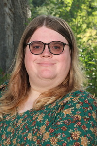 A person with long, wavy hair wearing sunglasses and a floral dress smiles in front of greenery and a tree.