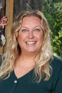 Smiling woman with long, wavy blonde hair, wearing a green top, stands outdoors with greenery in the background.